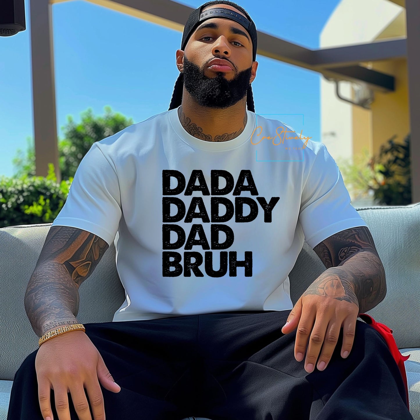 Young man sitting outdoors in a white T-shirt with bold black text: DADA, DADDY, DAD, BRUH. Wearing a backwards cap.