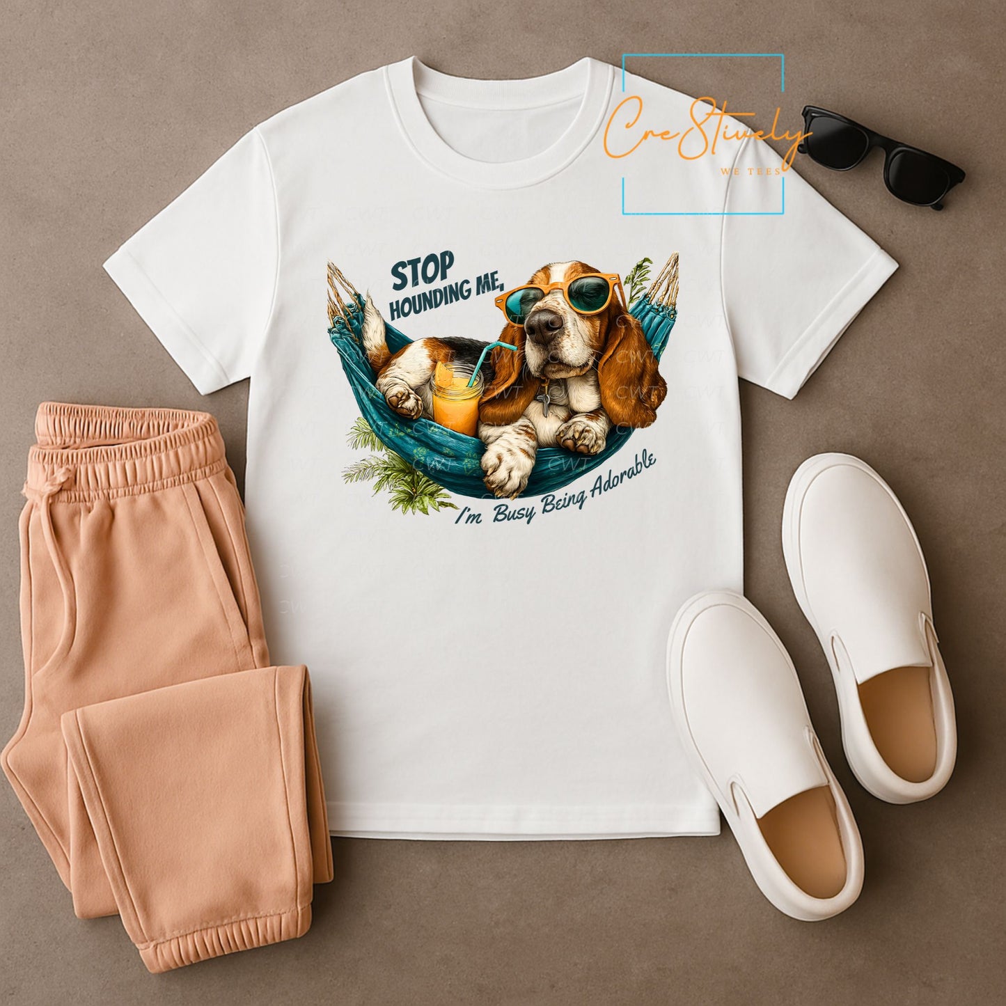 Flat lay of a white t-shirt featuring a cartoon hound dog in sunglasses lounging in a hammock with orange juice, reading “Stop Hounding Me, I’m Busy Being Adorable.”