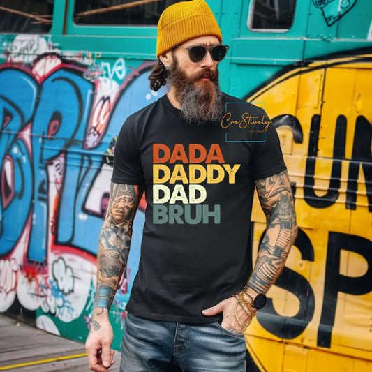 Bearded man in mustard hat wearing a black T-shirt with bold multicolored text: DADA, DADDY, DAD, BRUH. Standing against a graffiti wall.