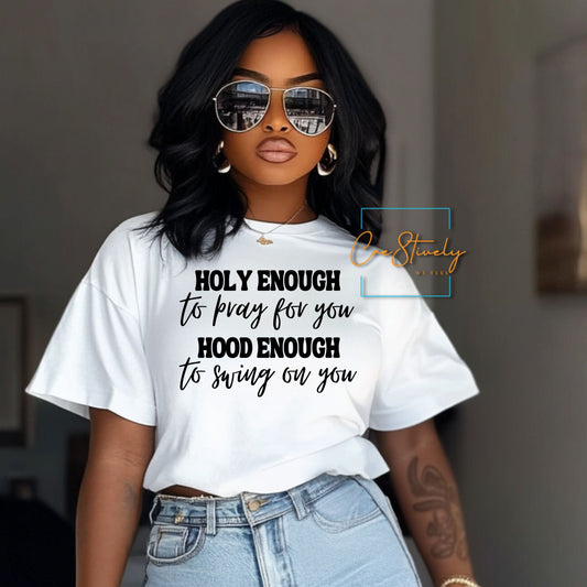 Woman wearing a white “Holy Enough to Pray, Hood Enough to Swing on You” tee, styled with sunglasses and high-waisted jeans, giving confident, cool-girl energy.