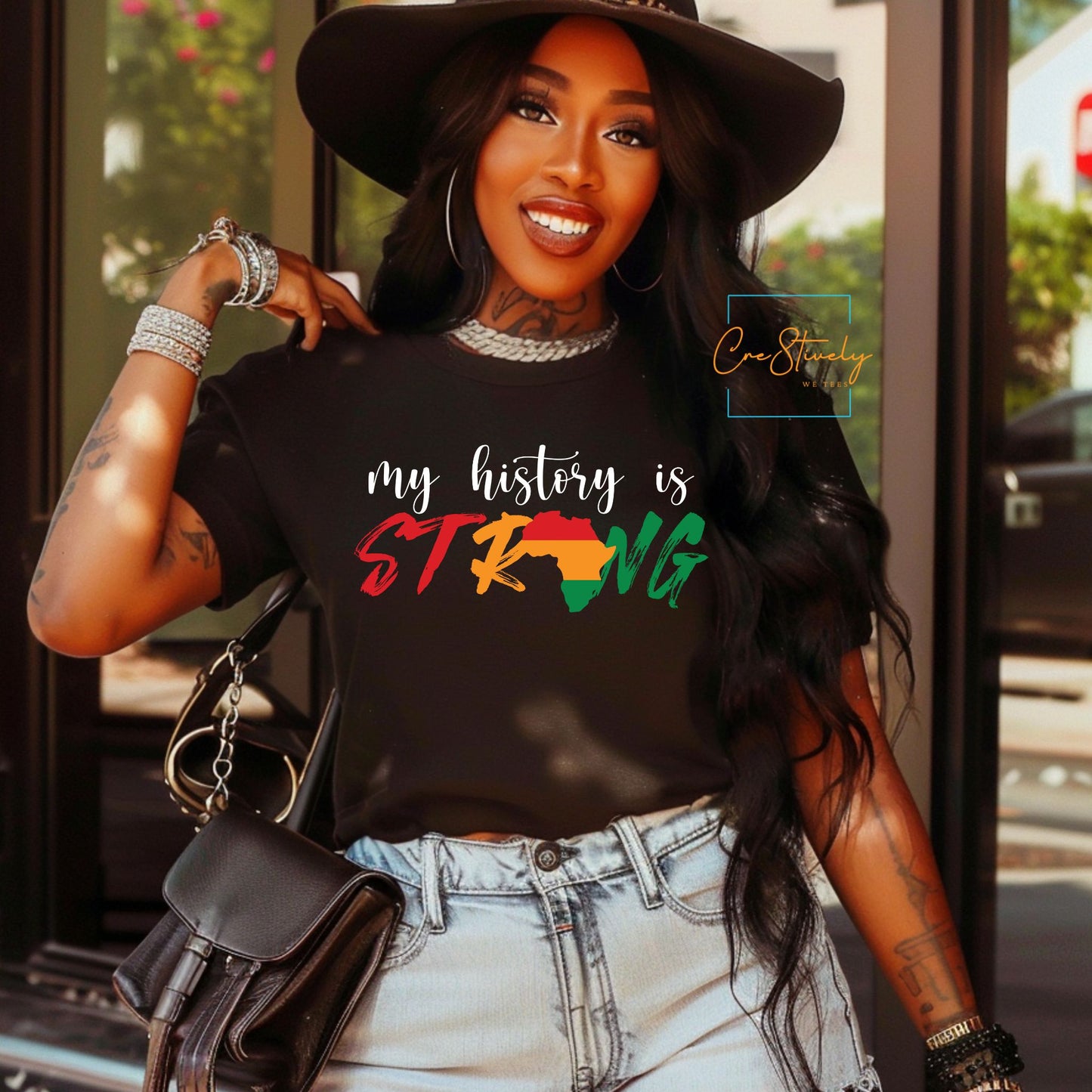 Black woman wearing a “My History Is Strong” t-shirt with Pan-African colors and an Africa-shaped letter "O," styled with a wide-brim hat, bold accessories, and a confident smile, promoting Black pride and cultural empowerment.