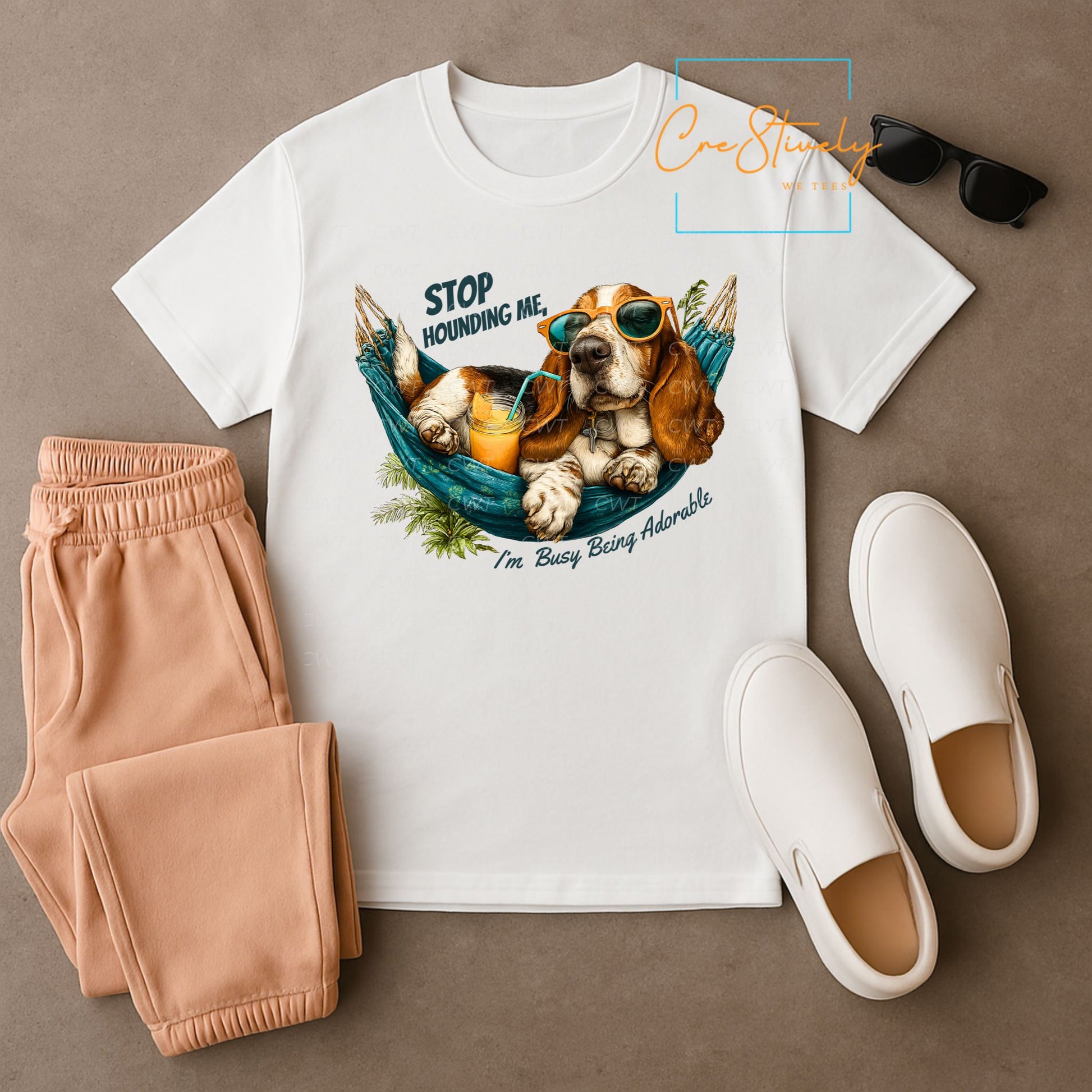 Flat lay of a white t-shirt featuring a cartoon hound dog in sunglasses lounging in a hammock with orange juice, reading “Stop Hounding Me, I’m Busy Being Adorable.”