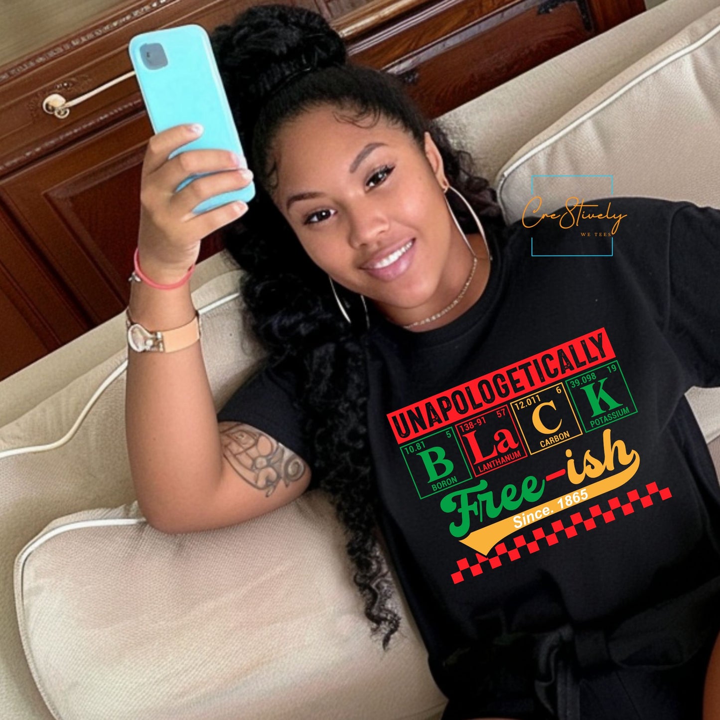 Woman lounging indoors in a black “Unapologetically Black, Free-ish Since 1865” t-shirt featuring a periodic table-inspired design in bold RBG colors, celebrating Black freedom and identity.